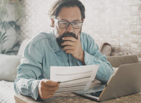 Mężczyzna siedzi przed laptopem i przegląda trzymany w jednej ręce papierowy dokument, drugą ręką gładząc brodę